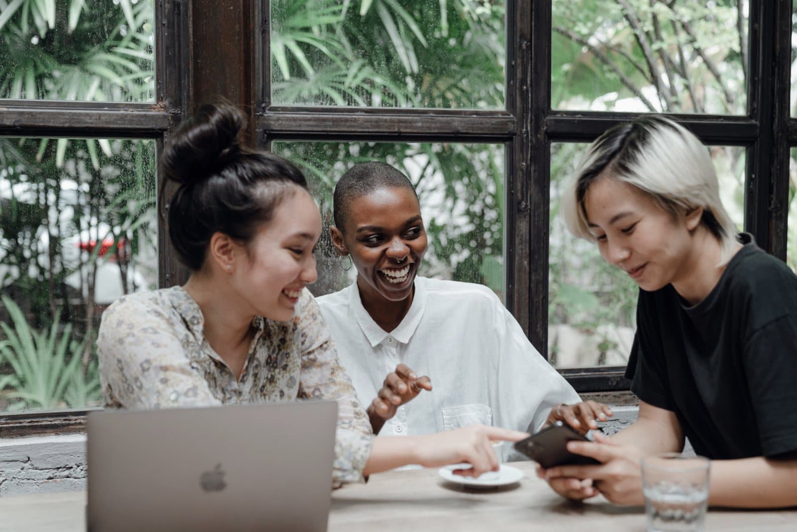 Three friends at a café table looking at a phone, with a laptop open